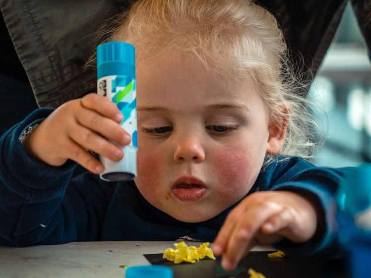Image of A young child with blonde hair focuses intently on gluing pieces of yellow material onto black paper, holding a glue stick in one hand. An adult stands close behind, assisting with the craft activity.