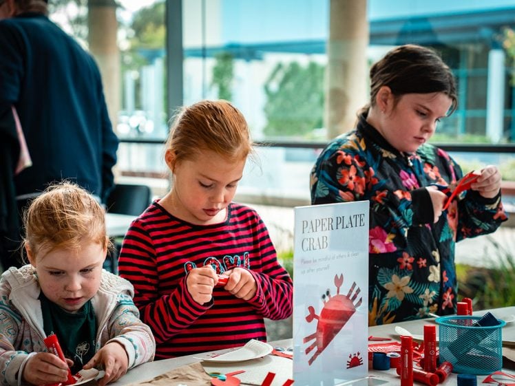 Image of Three young girls are focused on making crafts at a table. A sign reads Paper Plate Crab. Art supplies and finished crafts are on the table in a bright indoor space with large windows in the background.