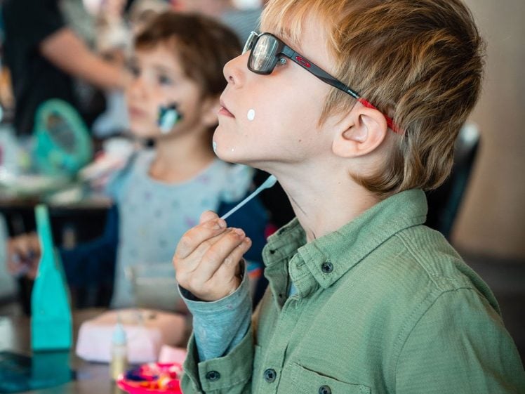 Image of A young boy wearing glasses and a green shirt applies white face paint to his chin with a brush, whilst other children participate in face painting activities at a busy indoor event.