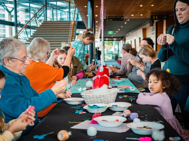 Image of A group of people of various ages sit around a long table, engaged in arts and crafts activities with wool and felt in a bright indoor space. A little girl in purple looks towards the camera.
