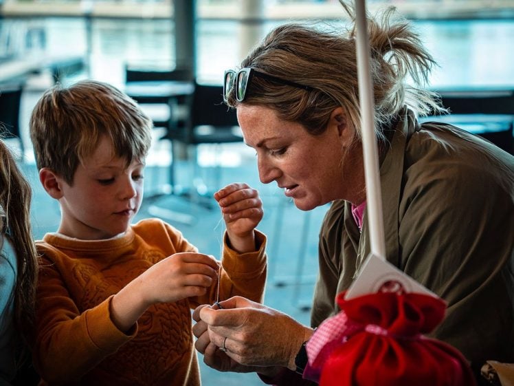 Image of A woman helps a young boy thread a needle, both focussed on the task. They sit at a table indoors, with a window and water visible in the background. The boy wears a mustard-coloured jumper.