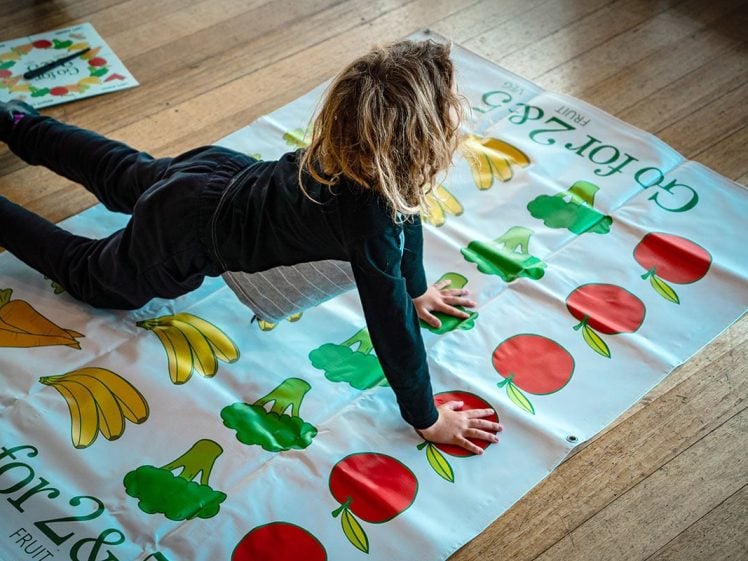 Image of A child with long, light brown hair does a plank on a colourful game mat with images of fruit and vegetables, including apples, bananas, and broccoli, on a wooden floor.