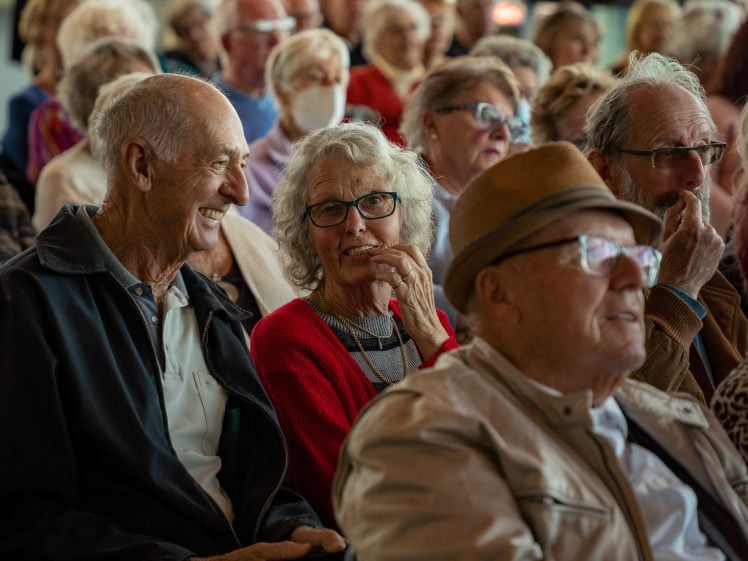 Image of A group of older adults sit closely together in an audience, attentively watching something. Many are smiling or engaged, some wearing glasses or hats, and one person in the background is wearing a face mask.