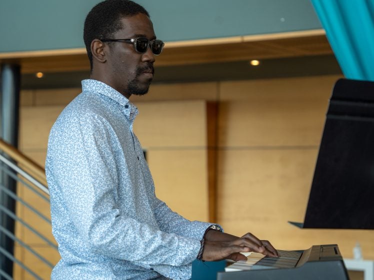 Image of A man wearing sunglasses and a light patterned shirt plays a keyboard piano indoors, with a wooden wall and staircase in the background.
