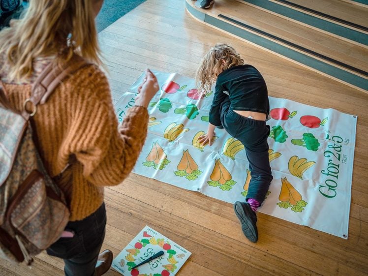 Image of A child plays a vegetable-themed floor game, placing hands and feet on images of carrots and other produce, whilst an adult watches nearby. The scene takes place indoors on wooden flooring.