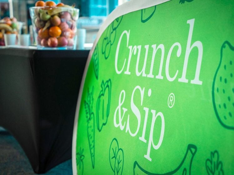 Image of A bright green banner reads Crunch & Sip with fruit illustrations. In the background, tables hold large glass bowls filled with assorted fresh fruit. The setting appears to be indoors.