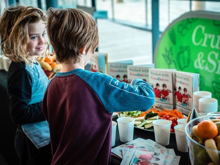 Image of Two children stand at a table with sliced vegetables, fruit, and cups. Several leaflets are displayed, and apples and oranges are in bowls. The children appear to be selecting snacks. A green sign in the background reads “Crunch & Sip.”.
