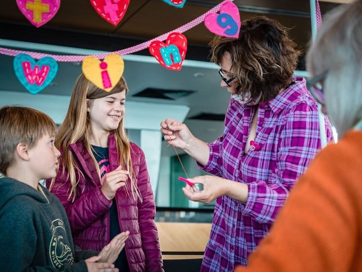 Image of A woman in a pink checked shirt helps two children with a craft project, threading string through a red heart shape. Colourful heart decorations hang above them in the background.