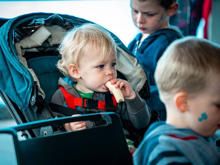 Image of A young child with blonde hair sits in a pushchair, eating a banana. Two other children, one with a blue sticker on his face, are nearby, all dressed in casual clothing. The setting appears to be indoors.