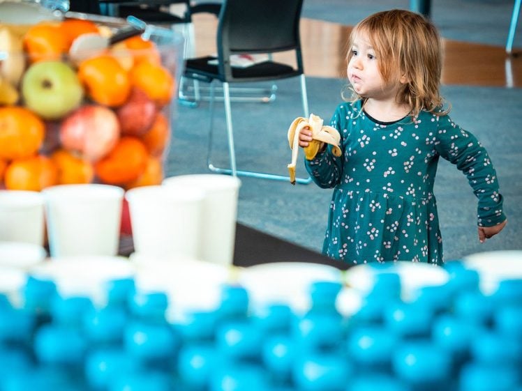 Image of A young child wearing a green dress with a floral pattern eats a banana whilst standing indoors near tables with cups, bottled water, and a bowl of assorted fruit.