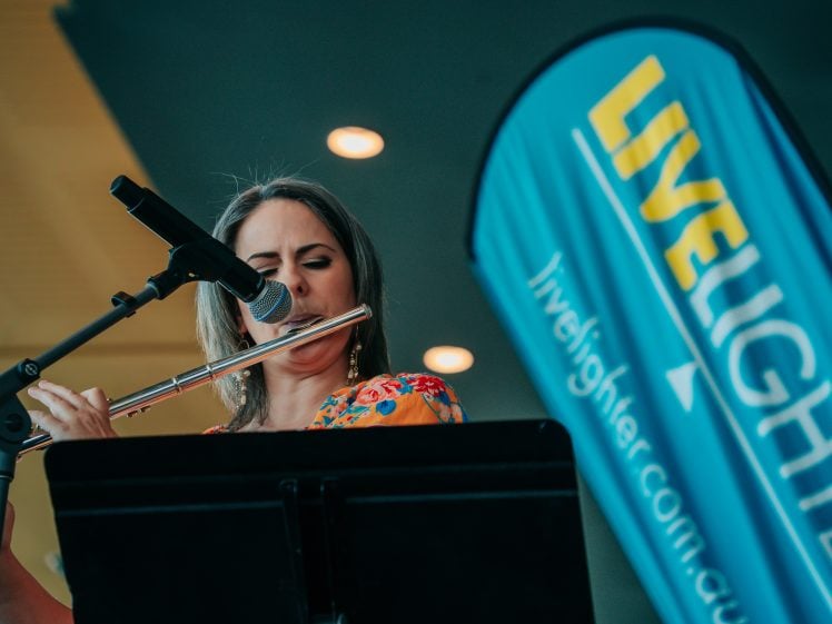 Image of A woman plays the flute and sings into a microphone. Next to her is a blue banner with the word LIVELIGHTER and the website livelighter.com.au.
