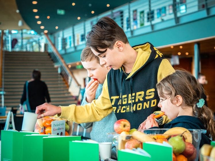 Image of Three children stand at a table with fruit and green leaflets, eating apples and oranges in a bright indoor setting with a staircase and bannister in the background.