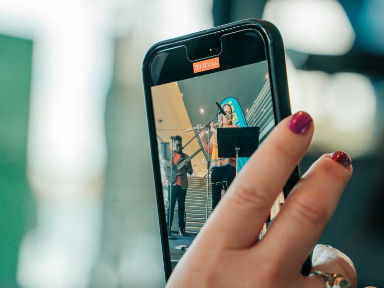 Image of A close-up of a hand holding a mobile phone, recording a video of two musicians performing indoors, one playing bass and the other singing into a microphone. The recording timer is visible on the phone screen.