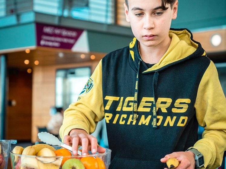 Image of A young person in a black and yellow Tigers Richmond hoodie selects fruit from a large clear bowl, with apples, oranges, tomatoes, and white paper cups on the table in front of them in a modern indoor setting.