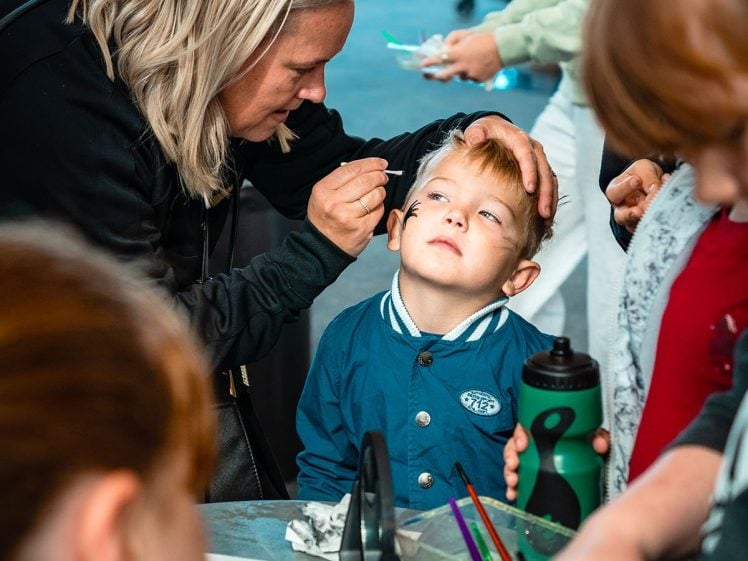 Image of A woman gently paints a young boy's face as he looks up, surrounded by children and art supplies on a table in a lively indoor setting.