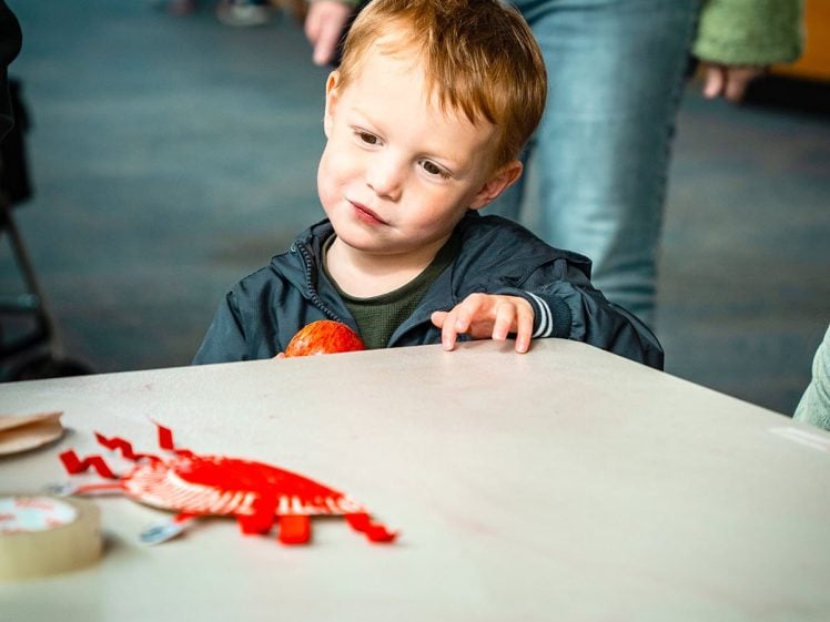 Image of A young boy with light brown hair looks thoughtfully at a table with craft materials, including red paper and tape, while an adult stands nearby in the background.
