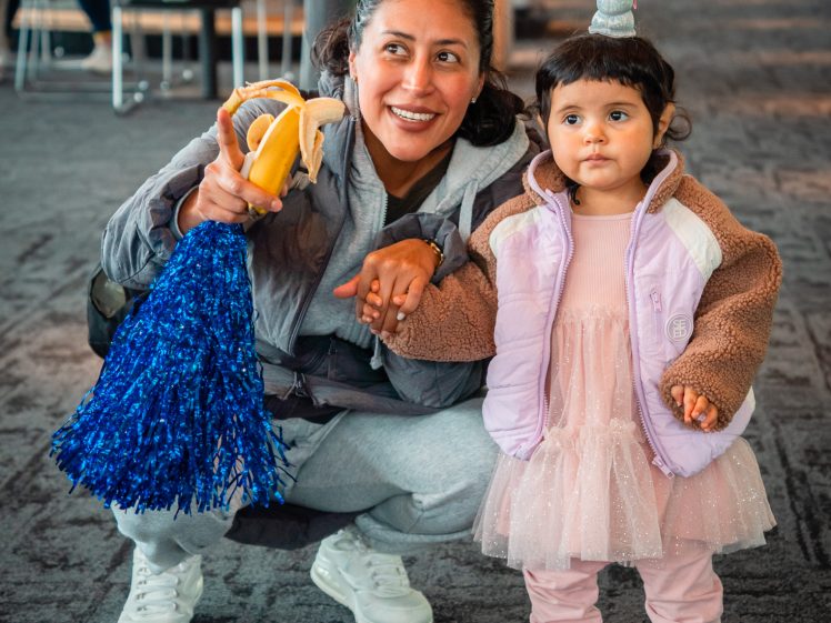 Image of A woman in sportswear squats, smiling and holding a banana and blue pom-pom, next to a young child in a pink dress and unicorn horn headband. They are indoors, holding hands, with people in the background.