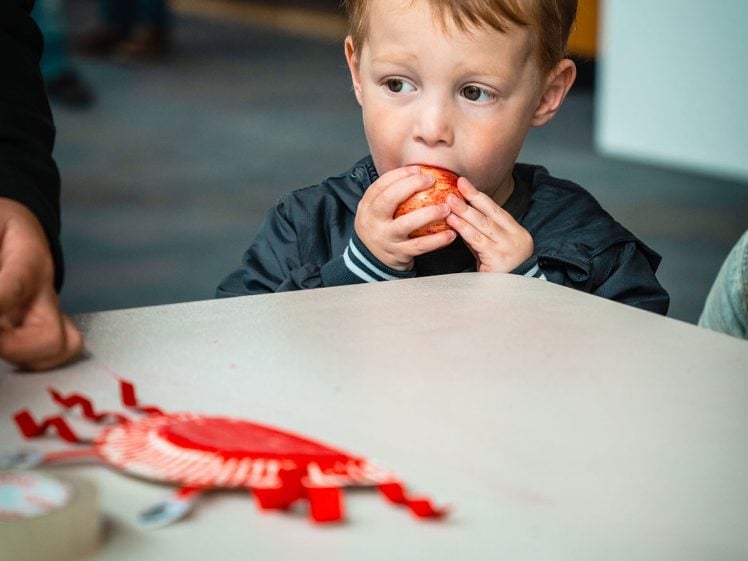 Image of A young child with light brown hair eats an apple whilst sitting at a white table with craft materials and red paper shapes on it. An adult’s hand is visible nearby. The background is out of focus.