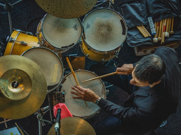 Image of A man in a black suit plays a yellow drum kit, seen from above. His drumsticks are poised over the drums, with cymbals and a case of additional drumsticks nearby.