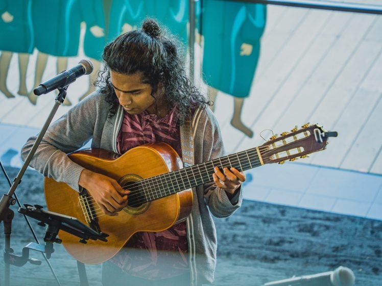 Image of A person with long curly hair plays an acoustic guitar and reads music on a stand, standing by a microphone in an indoor space with large windows and blue decor in the background.