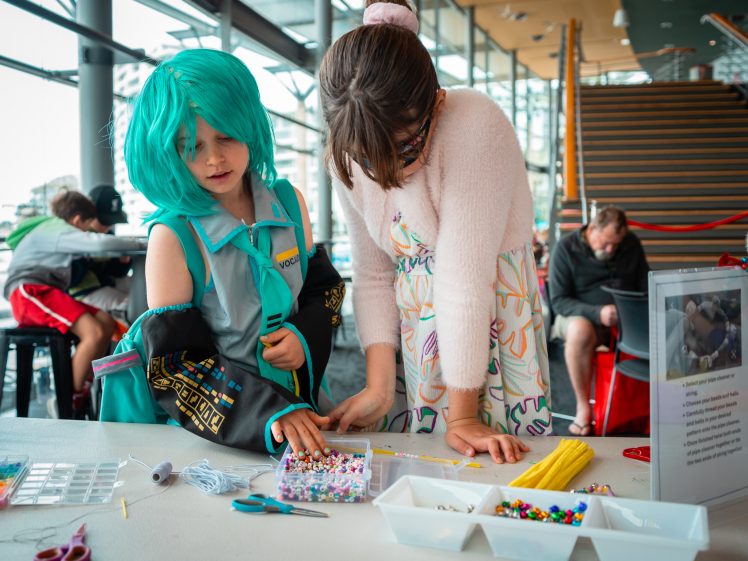 Image of Two children sit at a table making crafts with colourful beads and supplies. One child wears a blue wig and costume, while the other assists, both focused on their project in a bright, modern indoor space.