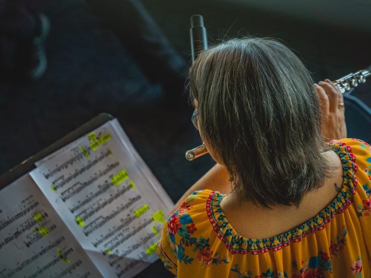 Image of A person with grey hair wearing a yellow floral top plays a woodwind instrument while reading sheet music on a stand, with highlighted notes visible on the pages. The scene is viewed from behind.