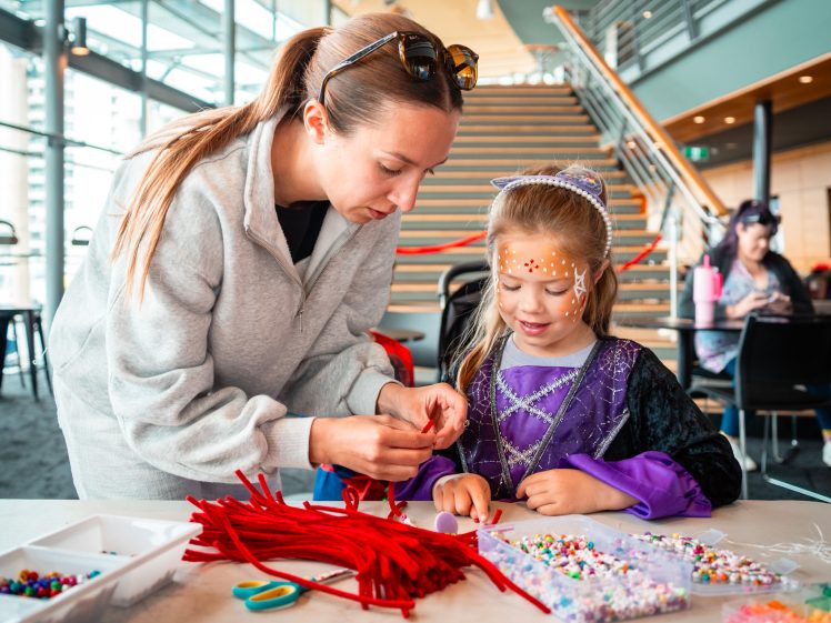 Image of A woman helps a young girl in a purple fancy dress decorate a craft at a table filled with beads and pipe cleaners in a bright, modern indoor space with stairs in the background.