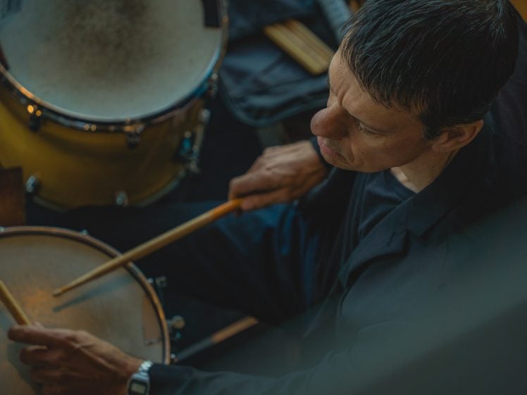 Image of An overhead view of a man playing drums, holding drumsticks and wearing a black shirt, with a drum kit partially visible in front of him.