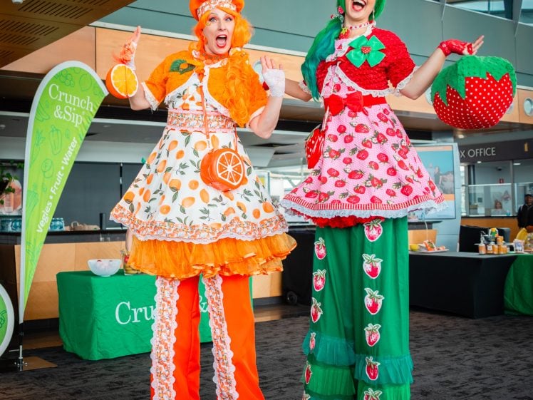 Image of Two performers on stilts wear colourful fruit-themed costumes—one in orange with citrus patterns and the other in red and green with strawberries. They pose cheerfully at an indoor event with banners in the background.