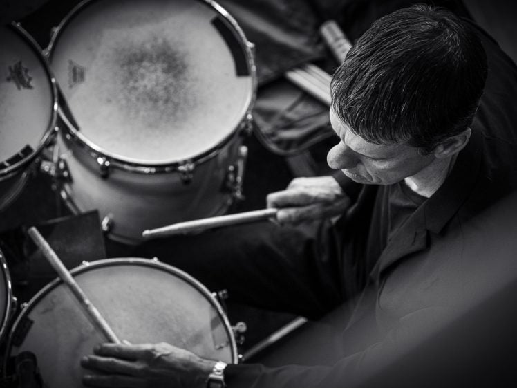 Image of Black and white photo of a man viewed from above, playing a drum kit with drumsticks in both hands, focussed on the drums in front of him.