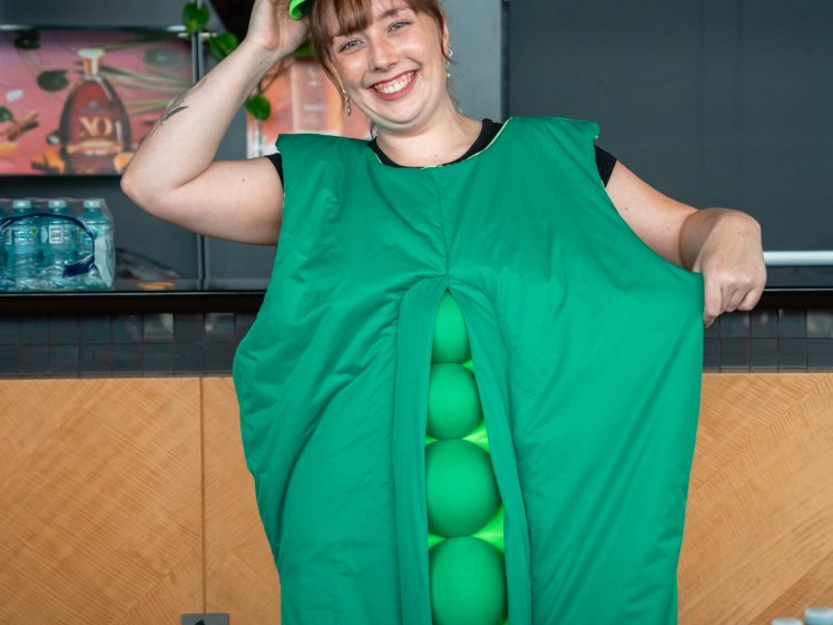Image of A smiling woman wearing a green pea pod costume and a pointed green hat stands indoors, holding her hat with one hand. In front of her, there is a table with bananas, a basket, and bottled water.