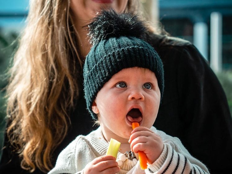 Image of A smiling woman with long hair sits behind a baby wearing a black knitted hat and striped jumper. The baby is eating vegetable sticks and has a beaded dummy clip around their neck.