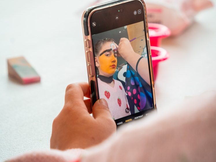 Image of A person holds a mobile, taking a photo of someone getting their face painted yellow with large black eyebrows. The mobile and painter’s hand are in focus; the background is blurred with art supplies scattered.