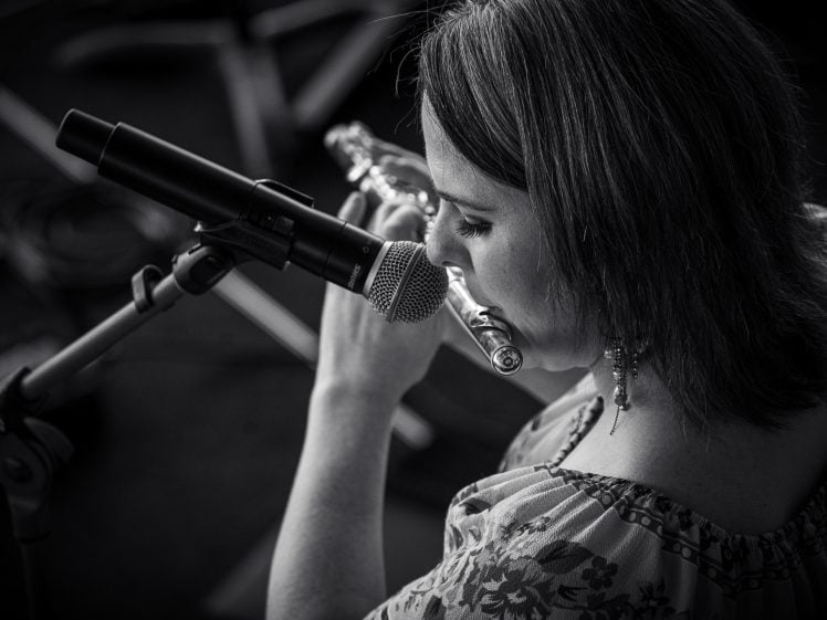 Image of A woman plays the flute into a microphone on a stand. The black and white photo is taken from above and slightly behind her, capturing her concentration as she performs.