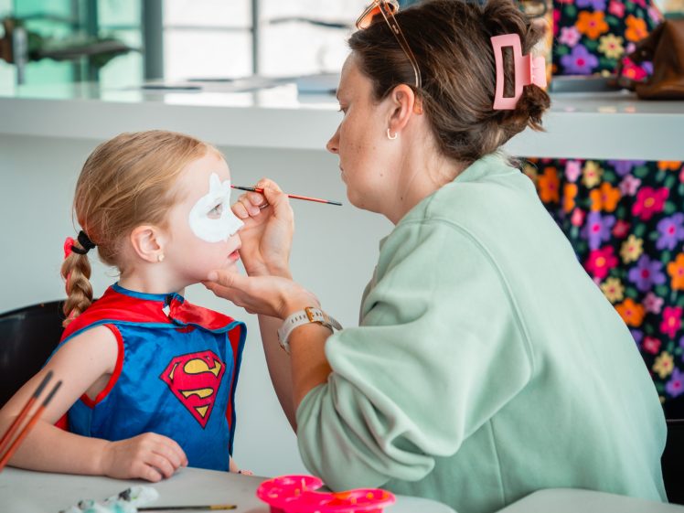 Image of A woman paints a white mask on a young girl’s face. The girl wears a blue Supergirl costume and red cape. They are seated at a table with face paints. The background is bright and colourful.
