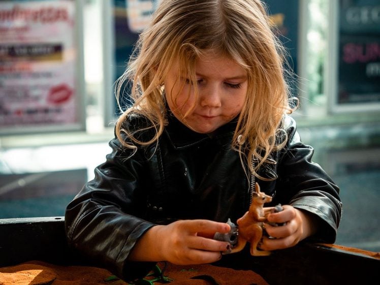 Image of A young child with long blonde hair, wearing a black jacket, plays with toy animals in a sandpit filled with orange sand. Colourful posters are visible in the blurry background.