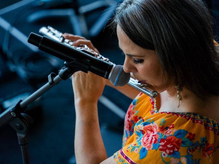Image of A woman in a colourful floral top plays the flute and sings into a microphone on stage, captured from above. Musical equipment and cables are visible in the background.