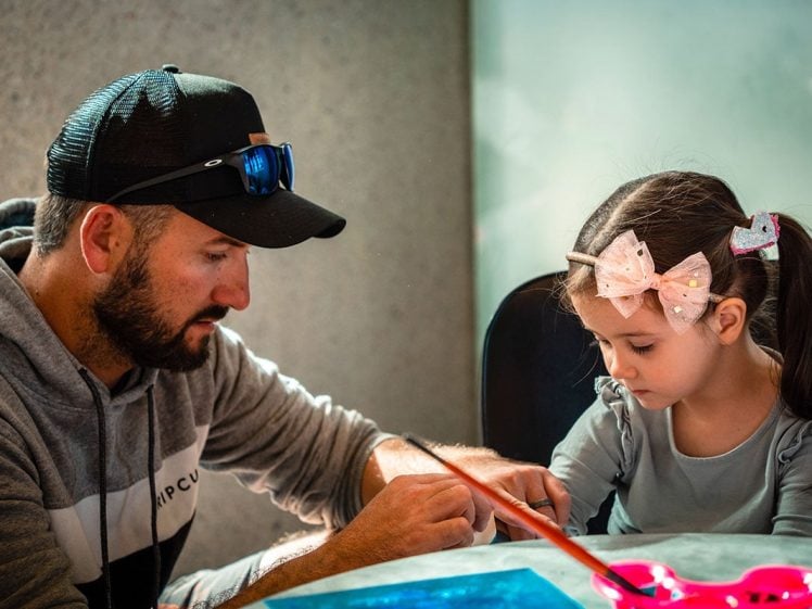 Image of A man wearing a cap helps a young girl with a pink bow in her hair as they sit at a table working on an art project together in a brightly lit room.