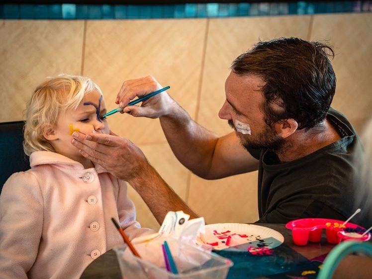 Image of A man paints a young child's face with blue and yellow designs while holding her chin gently. The child, wearing a light pink coat, sits patiently. Art supplies and paint are on the table in front of them.