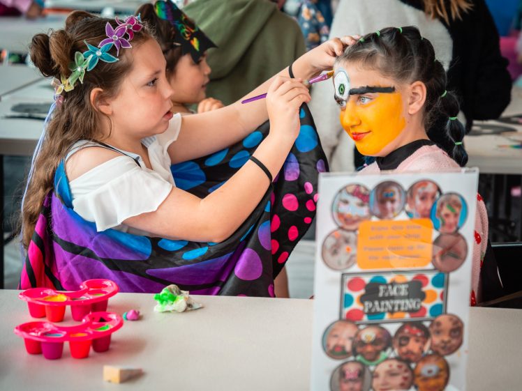 Image of Two children at a table, one painting the other's face with bright yellow and white colours. Art supplies and a face painting sign with sample designs are on the table. Other children are visible in the background.