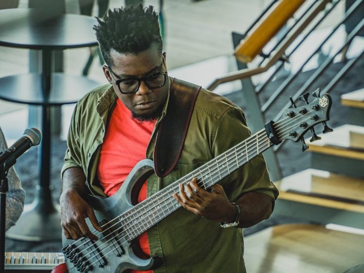 Image of A man wearing glasses and a red shirt plays a black electric bass guitar. He stands next to a microphone, with a staircase and tables in the background, focussing intently on his music.