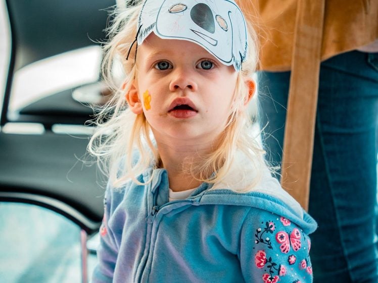 Image of A young child in a blue jacket with embroidered flowers wears a paper animal mask on their head and looks up with surprise. An adult stands nearby, holding a cup.
