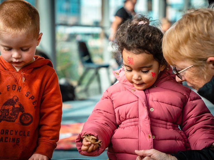 Image of Two young children and an older woman are engaged in an indoor activity. The girl in a pink coat holds a dried leaf, whilst the boy in an orange hoodie looks down. The background is softly blurred.