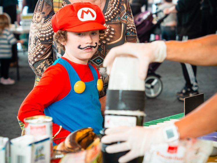 Image of A young child dressed as Mario from Super Mario stands with an older woman, watching a science demonstration involving bottles and various materials on a table at an indoor event.