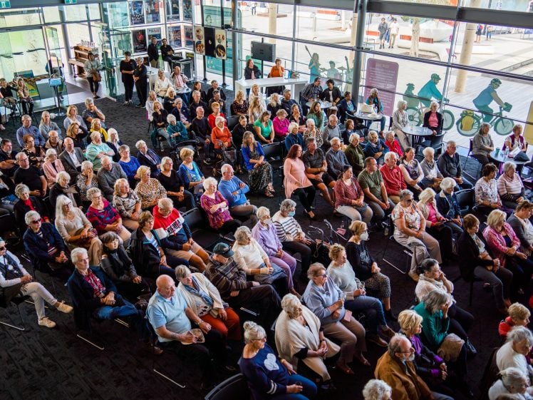 Image of A large group of people, mostly pensioners, are seated indoors, facing forward as if attending a presentation or event. The space is bright with floor-to-ceiling windows and some people standing at the back.