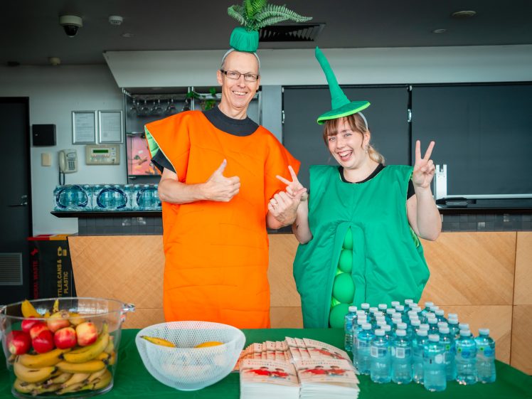 Image of Two people dressed in carrot and pea pod costumes stand behind a table with fruit, bottled water, and leaflets. They are smiling, giving a thumbs up and peace sign in a bright indoor setting.