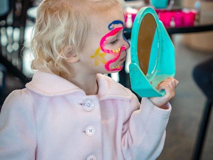 Image of A young child with colourful face paint looks into a small blue mirror whilst wearing a light pink coat. The child is indoors, and there are blurred people and tables in the background.