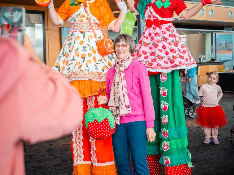 Image of Two performers on colourful stilts dressed as fruit-themed characters stand beside a smiling woman in pink glasses and a scarf, whilst a person takes their picture indoors. A child in a red tutu is visible in the background.