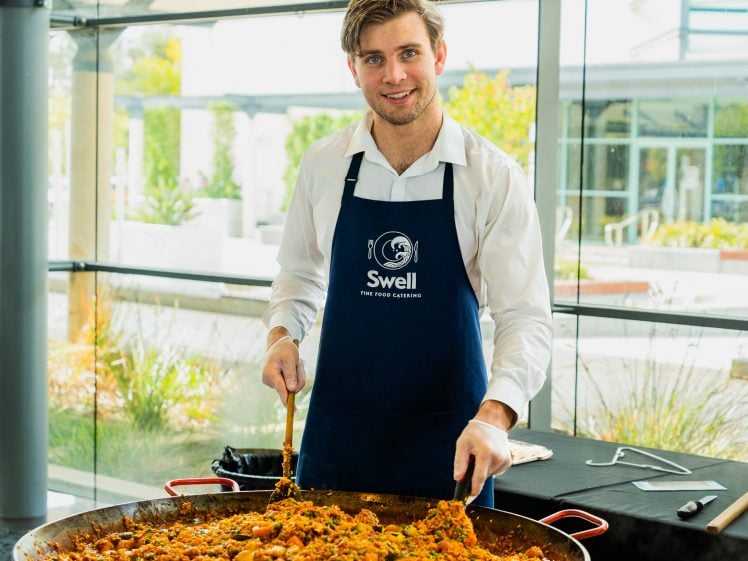 Image of A smiling man wearing a white shirt and a blue Swell Fine Food Catering apron serves a large pan of paella indoors, with glass windows showing greenery outside.