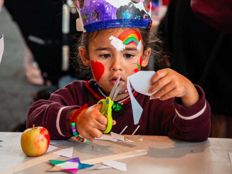 Image of A young child wearing a colourful paper crown and face paint uses green scissors to cut white paper at a table filled with craft supplies, with an apple and more paper nearby.